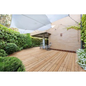 Taras z jasnobrązowych desek tarasowych t.b.t., zacieniony żaglem przeciwsłonecznym i otoczony zielenią.