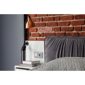 Zbliżenie sypialni z ceglaną ścianą, lampą i łóżkiem z szarym zagłówkiem.