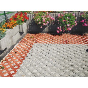 Ułożone kratki trawnikowe w kolorze szarym i czerwonym, obramowane szarym obrzeżem rabaty z kwiatami.