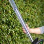 Akumulatorowe nożyce do żywopłotu LUX z akumulatorem i ładowarką na tle zielonego żywopłotu. Nożyce do żywopłotu z osłoną ostrza.
